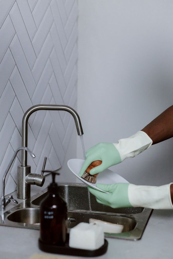 A person wearing green latex gloves washes a plate under running water in a modern kitchen sink.