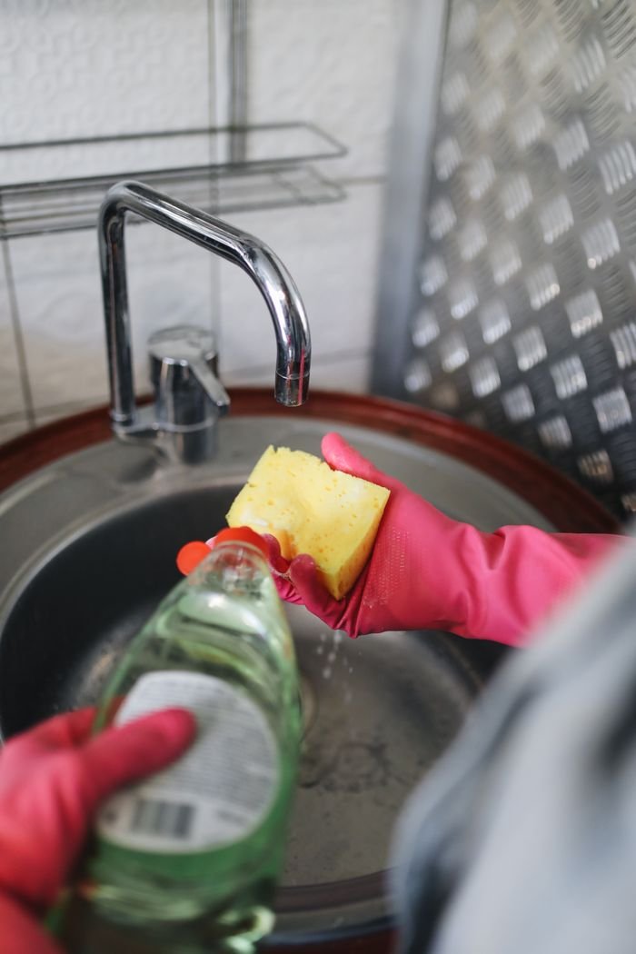 Hands wearing rubber gloves pouring detergent on a sponge over a kitchen sink.