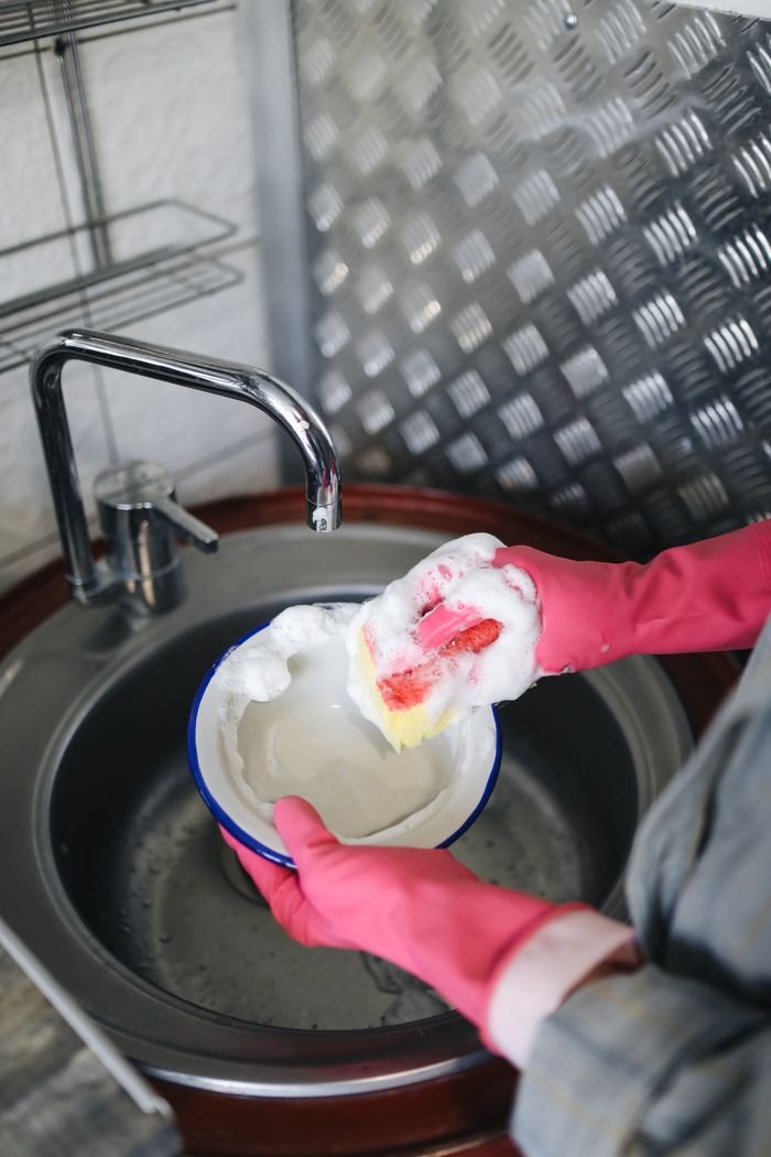 A person wearing gloves washes a dish with soap and foam over a stainless steel sink indoors.
