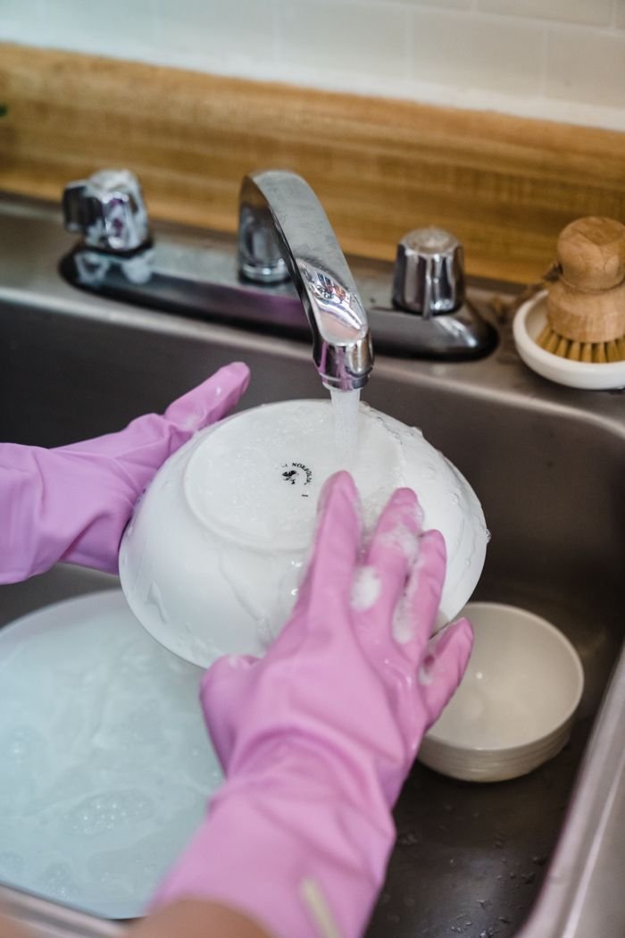 Hands wearing pink gloves washing dishes in a kitchen sink with running water.